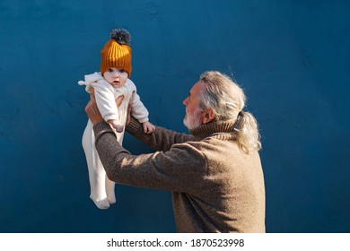 Senior Man Holding Little Baby. Side View Of Happy Gray Haired Grandfather Holding Adorable Little Baby In Outstretched Hands Against Blue Background