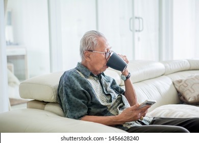 Senior Man Holding A Mobile Phone While Drinking A Cup Of Hot Tea And Sitting On The Couch