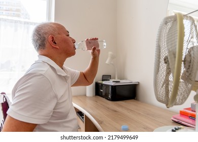 A Senior Man At Home Drinking Water Next To A Electric Fan On A Record Breaking Hot Summer Day Trying To Avoid Dehydration And Heat Stroke.