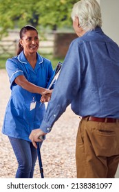 Senior Man At Home Using Walking Stick Greeting Female Nurse Or Care Worker In Uniform At Door