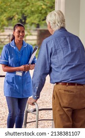 Senior Man At Home Using Walking Frame Greeting Female Nurse Or Care Worker In Uniform At Door