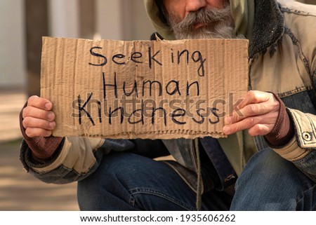 Senior man homeless beggar with a gray beard in a shabby clothes with a carboard sign sitting outdoors in city and asking for money donation. Sign on paperboard Seeking Human Kindness