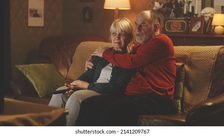 Senior Man Hugging Crying Woman While Resting On Sofa And Watching Sad Movie On TV Or Evening News, In Evening In Cozy Living Room At Home