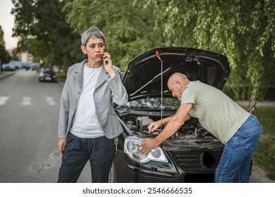 A senior man husband inspect a car engine with an open hood while a woman in a blazer makes a phone call on a quiet street, likely seeking roadside assistance. - Powered by Shutterstock