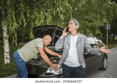 A senior man husband inspect a car engine with an open hood while a woman in a blazer makes a phone call on a quiet street, likely seeking roadside assistance. - Powered by Shutterstock