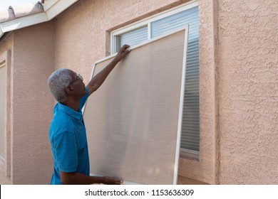  A Senior Man Installing A Window Screen On House For Summer Or Spring Seasons.