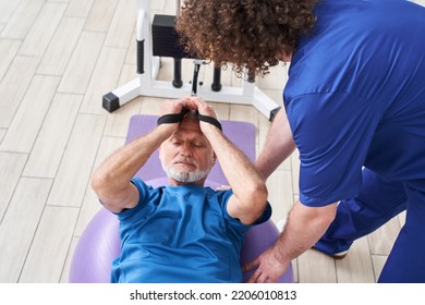 Senior Man Laying On A Fitness Ball In Physical Rehabilitation Therapy With His Trainer