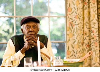 Senior man leans on his elbows and clasps his hands as he sits at a dining room table and poses for a portrait.