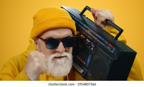 Senior Man Listening Record Player On Yellow Studio Background. Cheerful Old Guy Dancing With Player On Shoulder Indoors. Joyful Male Person Enjoying Retro Music Inside.