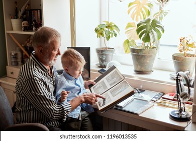 Senior Man And Little Boy Holding And Looking At Family Photo Album In Living Room