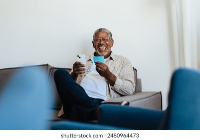 Senior man looking confident while shopping online using his smartphone and credit card. Happy older adult enjoying online banking from the comfort of his home. - Powered by Shutterstock