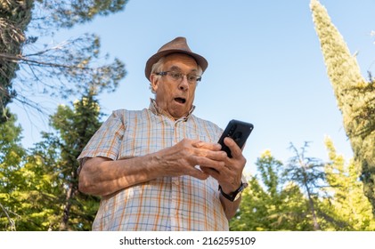 Senior Man Looking At His Mobile Phone With A Surprised Expression In The Park. Horizontal Photography, With Space For Text.