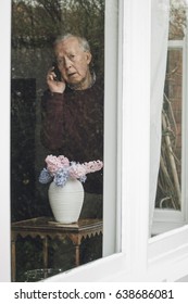 Senior Man Is Looking Out From His Window Into The Street And Is Reporting A Neighborhood Crime On The Phone.