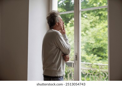 Senior man looking out window with hand on face, standing in a bright room, capturing a thoughtful and reflective moment, highlighting introspection and peacefulness in a serene setting - Powered by Shutterstock