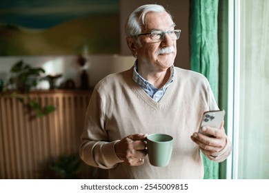 Senior man looking out window holding smartphone and coffee mug - Powered by Shutterstock