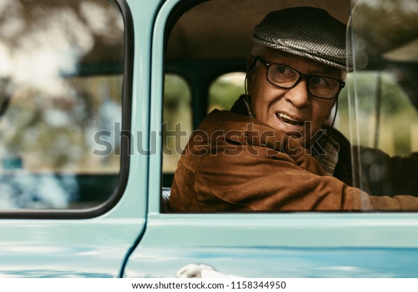 Senior man\
looking out of window while driving car. Elderly man wearing cap\
and eyeglasses driving a vintage\
car.