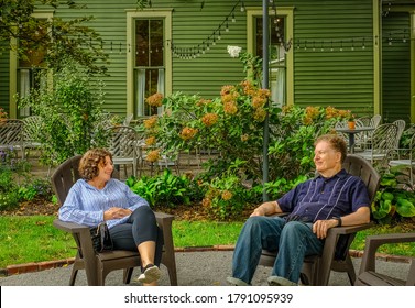 Senior Man And Mature Woman Sitting On Lounge Chairs N Front Of House Talking And Smiling; Fall In Midwest