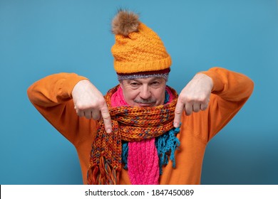 Senior Man With Mustache Wearing Several Scarfs And Hats Pointing To The Side To Present A Product. Studio Shot On Blue Wall.