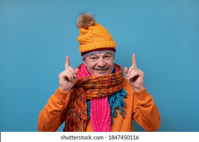 Senior Man With Mustache Wearing Several Scarfs And Hats Pointing To The Side To Present A Product. Studio Shot On Blue Wall.