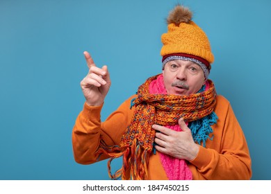 Senior Man With Mustache Wearing Several Scarfs And Hats Pointing To The Side To Present A Product. Studio Shot On Blue Wall.