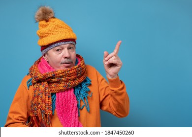 Senior Man With Mustache Wearing Several Scarfs And Hats Pointing To The Side To Present A Product. Studio Shot On Blue Wall.