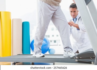 Senior Man On Treadmill With Therapist Crouching In Fitness Studio