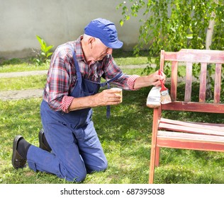 Senior Man In Overalls Painting Old Bench In Garden After Sandblasting. Repairing Old Furniture