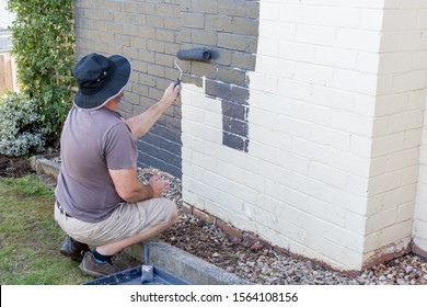 Senior Man Painting The Exterior Of A Brick Suburban Home 