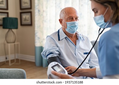 Senior man in pajamas getting blood pressure checked from nurse with face mask for safety against covid19 virus. Nurse checking blood pressure of old patient wearing safety precautions against Covid.