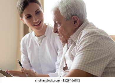 Senior Man Patient And Young Woman Caregiver Medical Worker In Uniform Hold Clipboard Noting Personal Information Talking Listens Client Telling About Health Complaints, Care Support Nursing Concept