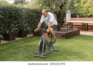 A senior man pedals on a stationary bike in a green garden setting, promoting fitness and outdoor activity. Day time, sunlight. Horizontal, wide angle, copy-space. - Powered by Shutterstock