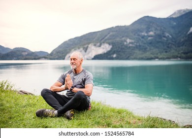 A Senior Man Pensioner Sitting By Lake In Nature, Doing Yoga Exercise.