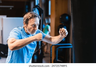 A senior man performs martial arts by punching a bag in a gym. He is wearing a blue T-shirt and is very focused on his training. - Powered by Shutterstock