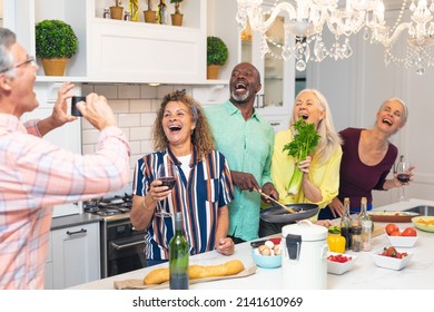 Senior man photographing laughing multiracial male and female friends cooking food at home. unaltered, lifestyle, cooking, preparation, social gathering, domestic life and togetherness.