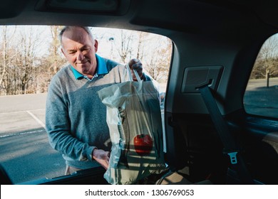 Senior Man Is Placing Shopping Bags In The Boot Of His Car. 