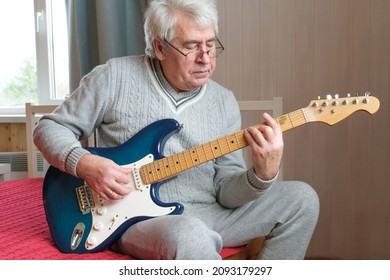 Senior man is playing guitar. Elderly man sitting on the sofa and playing guitar. Portrait of a gray-haired mature man in a sweater and glasses learning to play. Enjoying retirement life at home.