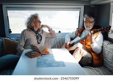 Senior man is playing guitar for his smiling wife who is reading a book inside their camper van, during a relaxing holiday trip - Powered by Shutterstock