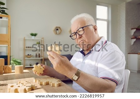 Senior man playing with jigsaw parts. Old man with Alzheimer's disease sitting at desk in geriatric clinic or nursing home, playing game, looking at wood puzzle piece he's holding in hand and thinking
