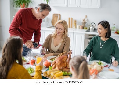 Senior Man Pouring Wine Near Smiling Multicultural Family During Thanksgiving Dinner At Home