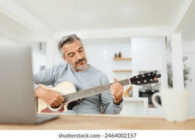 Senior man practicing guitar chords, engaged and thoughtful in a stylishly decorated kitchen with laptop. - Powered by Shutterstock