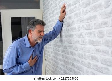 Senior man presses hand to chest has heart attack suffers from unbearable pain closes eyes wears  poses against grey brick wall background. People age and problems with health concept, copy space.