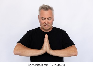 Senior Man Pressing Hands Together. Male Model In Black T-shirt Clasping Hands, Standing With Closed Eyes, Praying Or Meditating. Portrait, Studio Shot, Meditation Concept