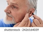 Senior man putting a hearing aid on his ear while visit his doctor at hearing clinic. Hearing solutions for elderly deafness people