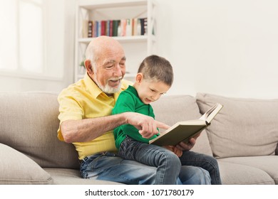Senior man reading book for his grandchild. Cheerful grandfather telling fairy tale aloud to his excited grandson, sitting on couch at home, copy space