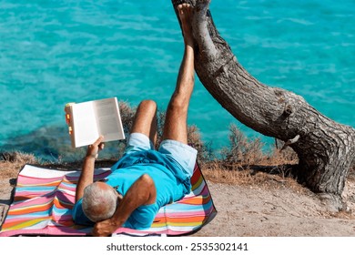 Senior man reading a book on the beach leaning on a tree and enjoying his vacation - Powered by Shutterstock