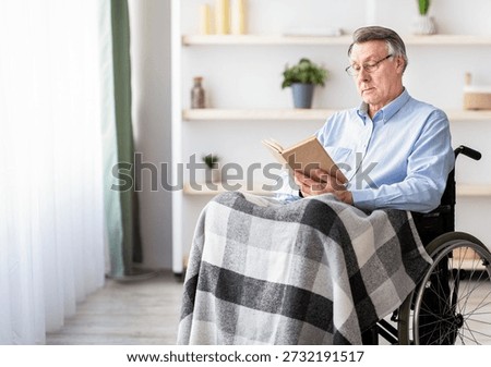 Senior Man Reading a Book While Sitting in a Wheelchair Covered With a Blanket Indoors
