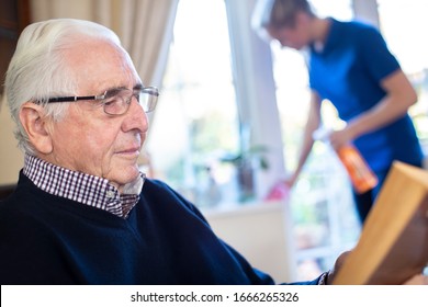 Senior Man Reading In Foreground Whilst Female Home Help Cleans House 