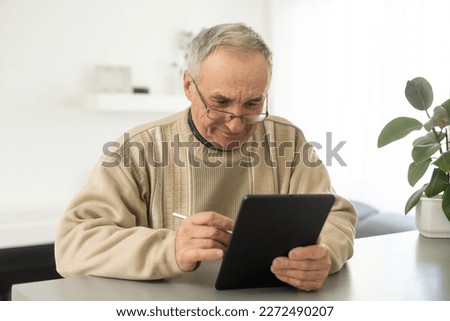 Senior man reading news on digital tablet. Serious mature male using portable computer at home, sitting at table, copy space.