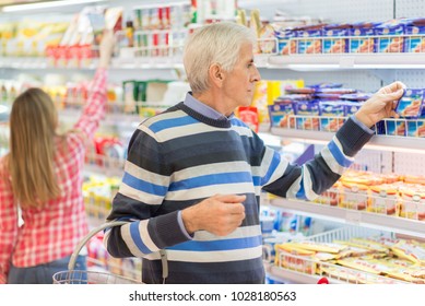 Senior Man Reads The Nutrition Label On A Package Of Food In Grocery Store.A Woman Is Shopping In The Background.