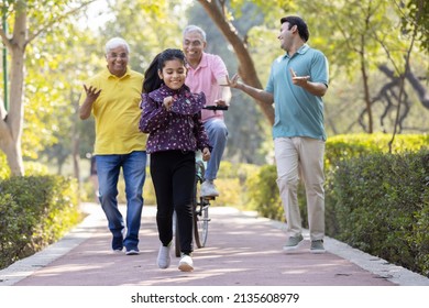 Senior Man Riding Bicycle With Granddaughter Running While Other Old Man And Son Encouraging At Park
